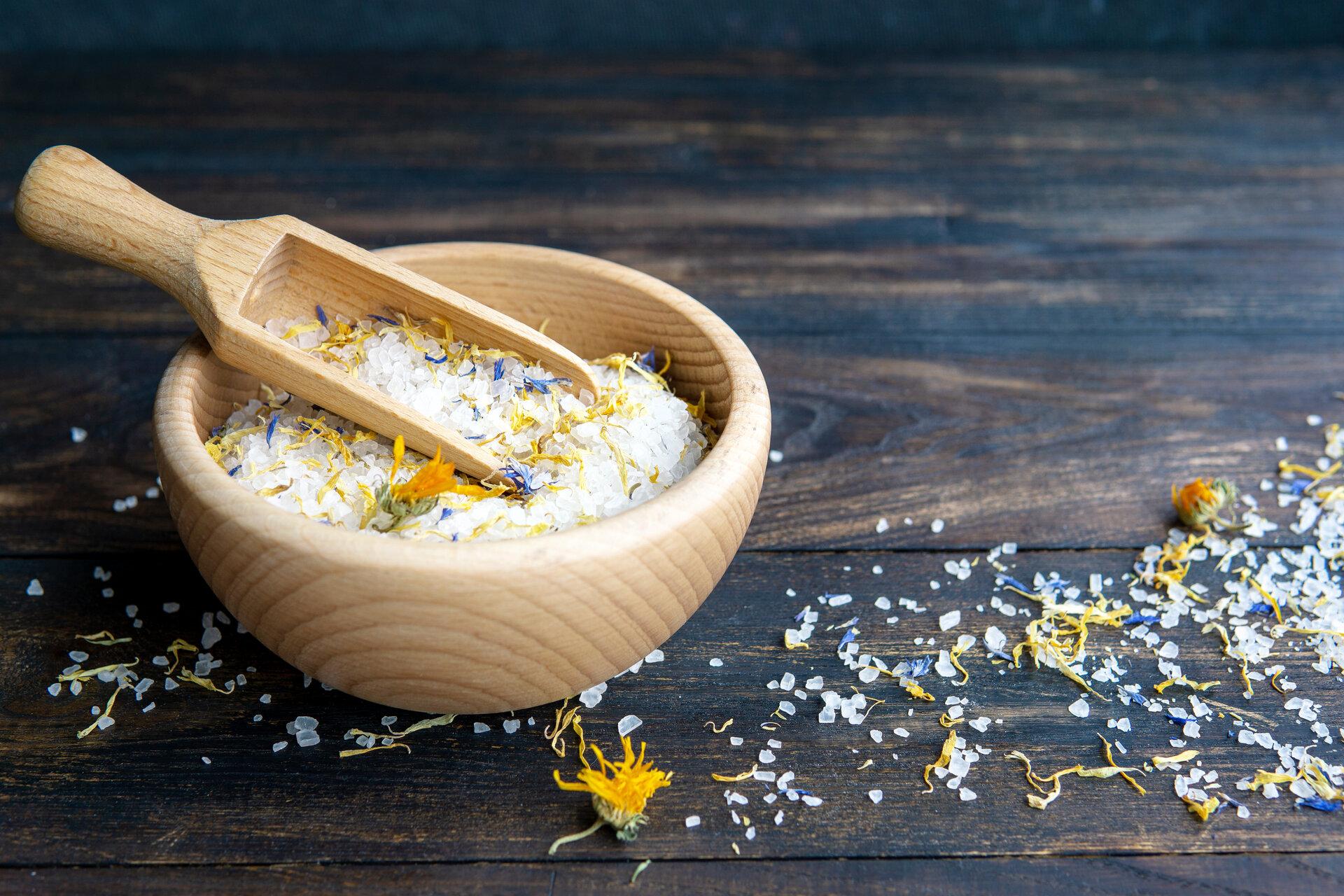 White fragrant sea salt crystals with dried calendula flowers in wooden bowl with spoon on dark rustic wooden background. Spa, body and health care concept.